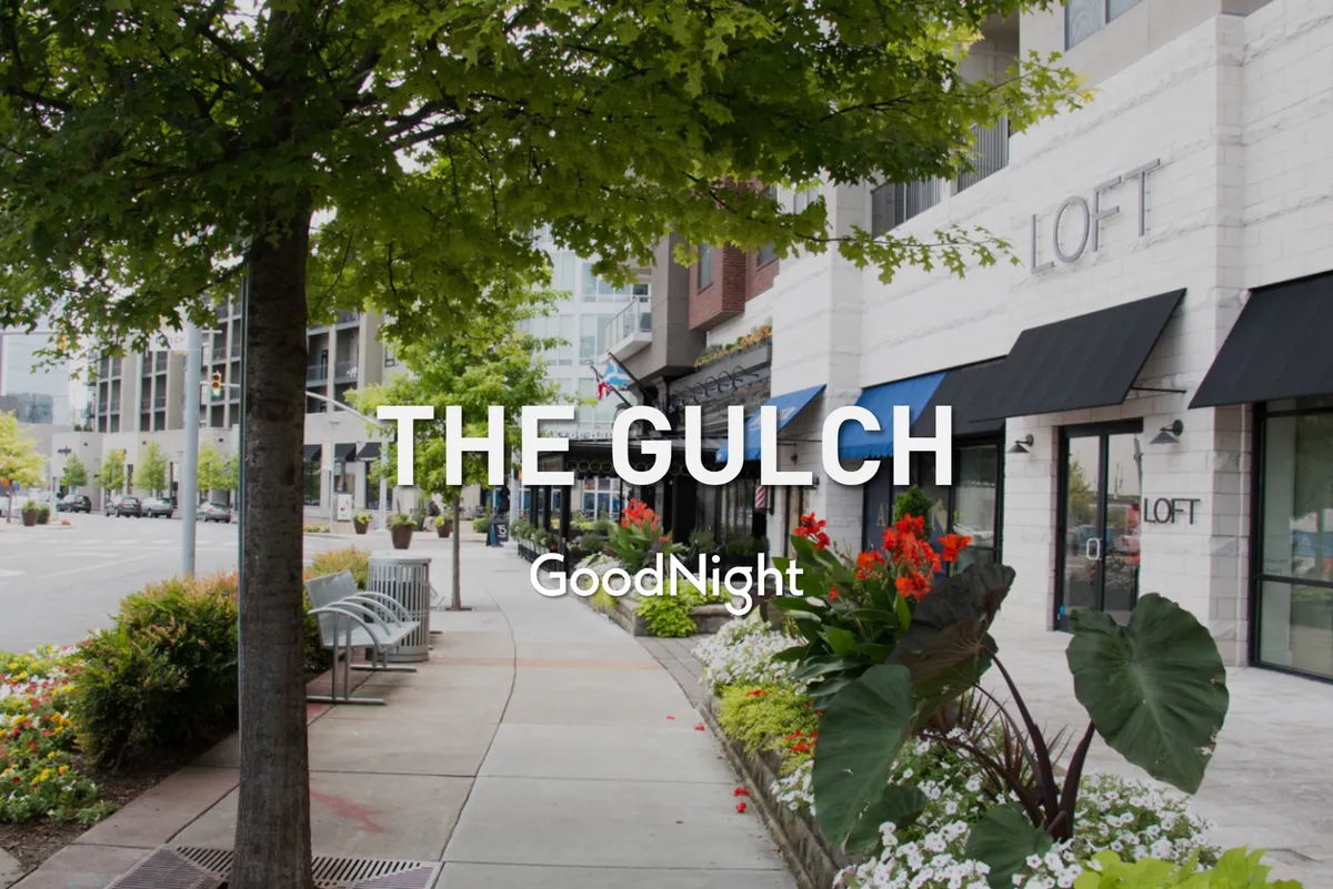 A charming streetscape lined with storefronts, flower beds, benches, and a large tree, evoking the feel of a walkable, community-oriented neighborhood. The scene captures people strolling, shopping, and enjoying the local ambiance, echoing the boutique and artisan shop experience described in the section.