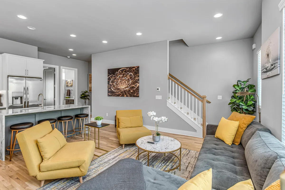 This image captures an inviting living area featuring a bright yellow armchair, a stylish gray sofa, a modern coffee table, and artwork on the wall. The kitchen area is visible in the background, showcasing the open flow between the living and culinary spaces. Natural light fills the space, highlighting its cheerful, social atmosphere.