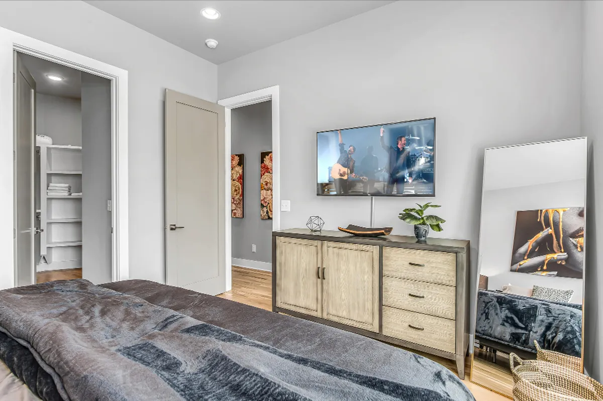 A comfortable bedroom setup with a queen bed, television mounted above a sleek dresser, and a mirror reflecting soft lighting. The room exudes a cozy and welcoming vibe, ideal for relaxing or enjoying quiet entertainment.