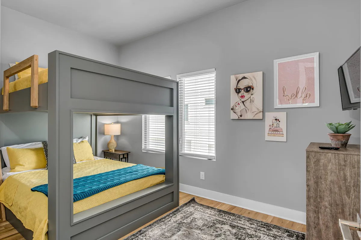 A cheerful bedroom with bright yellow bedding on a queen bunk bed, whimsical artwork on the walls, a sturdy wooden dresser, and windows with blinds letting in daylight. The setup is both fun and practical, perfect for families or groups.