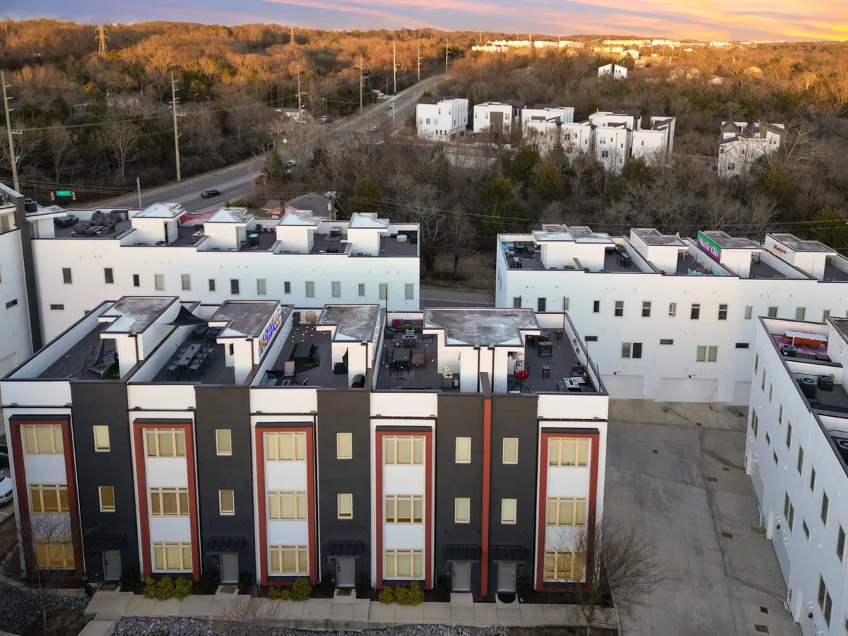 Rows of modern townhouses with private rooftop terraces are set against a sophisticated cityscape and surrounding greenery. The rooftops hint at social and informal meeting spaces, while the neighborhood setting underscores accessibility and comfort.