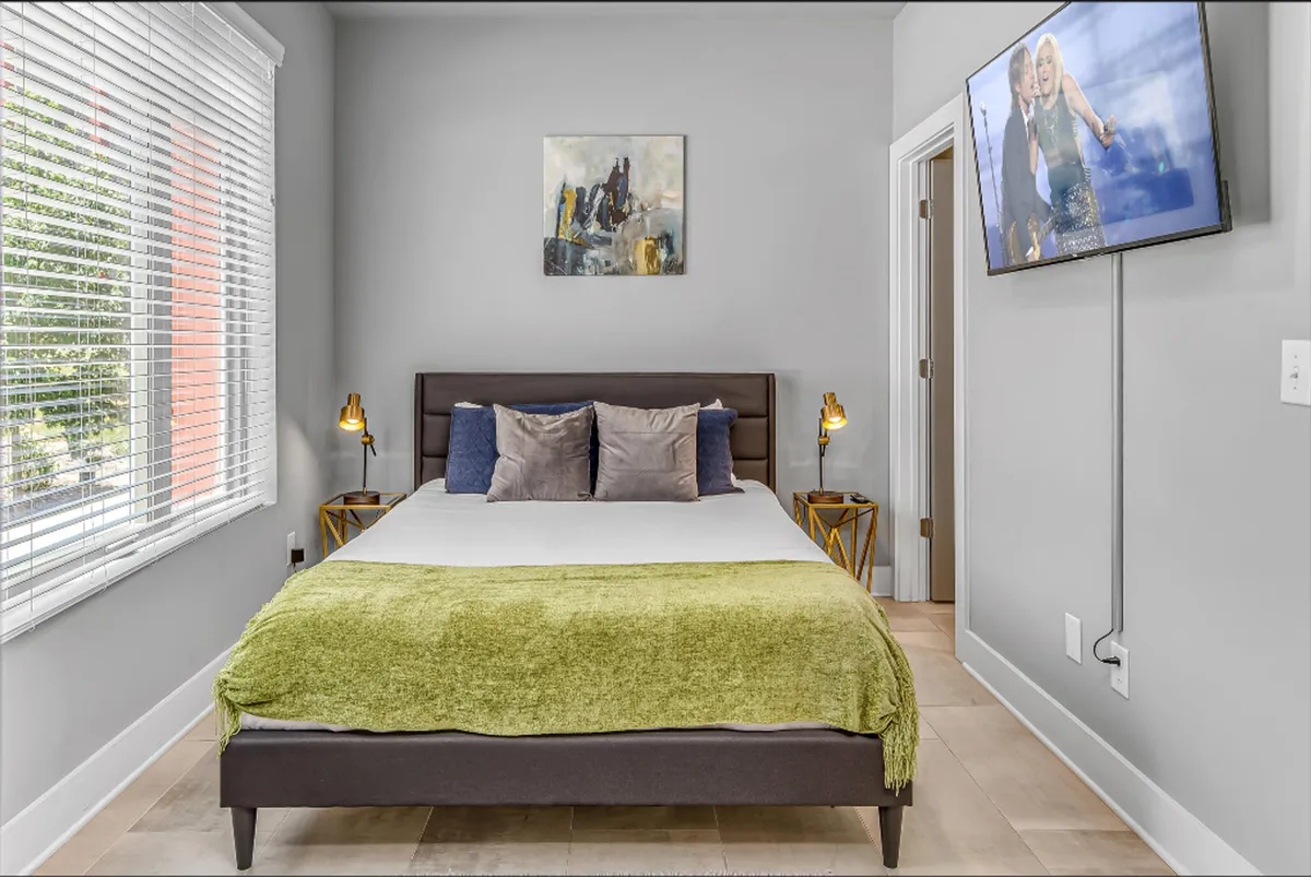 A tranquil bedroom featuring a plush bed, stylish artwork above the headboard, bedside lamps providing soft lighting, and a wall-mounted smart TV. Natural light filters in, enhancing the serene and restful atmosphere ideal for relaxation or a cozy movie night.