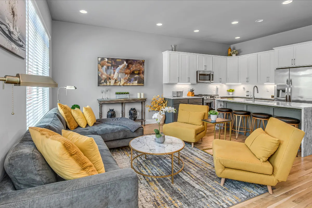 The image showcases a bright, inviting open-concept living space featuring vibrant yellow chairs, a modern coffee table, artistic wall décor, and a spacious kitchen area. The setting is designed for entertaining, relaxing, and spending quality time with friends or family, capturing a cheerful, welcoming atmosphere.