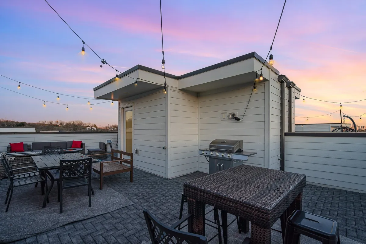 A stylish rooftop patio illuminated by warm string lights as the sun sets. The space features a modern grill, inviting outdoor seating, and a vibrant sunset sky in the background, suggesting a lively, sociable atmosphere ideal for gatherings and relaxation.