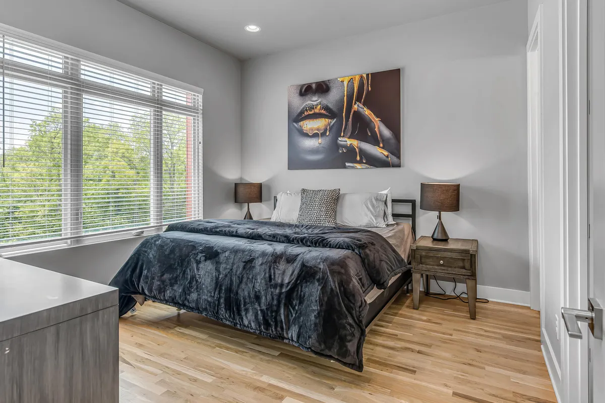 A serene bedroom featuring a plush bed with a soft blanket, modern artwork on the wall, and nightstands with lamps on either side. The space is filled with natural light, creating a relaxed and contemporary ambiance ideal for rest.