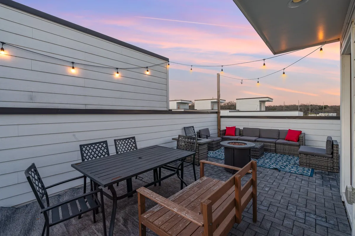 A cozy rooftop setting with string lights overhead, a fire pit surrounded by plush outdoor seating, and a long dining table set for an alfresco meal. The scene captures a welcoming ambiance, ideal for both large celebrations and intimate gatherings.