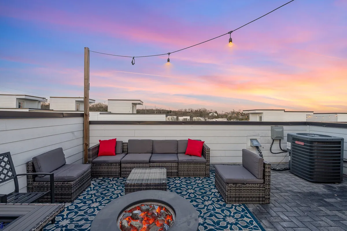 A stylish outdoor rooftop setting with a fire pit, comfortable seating, string lights, and a patterned rug. The exterior atmosphere is cozy and ideal for evening gatherings, with city views in the background.