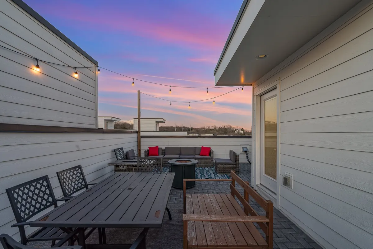 A cozy rooftop outdoor seating area adorned with string lights overlooking a sunset sky. The space features a fire pit and comfortable seating, perfect for evening gatherings, laughter, and toasts under the stars.