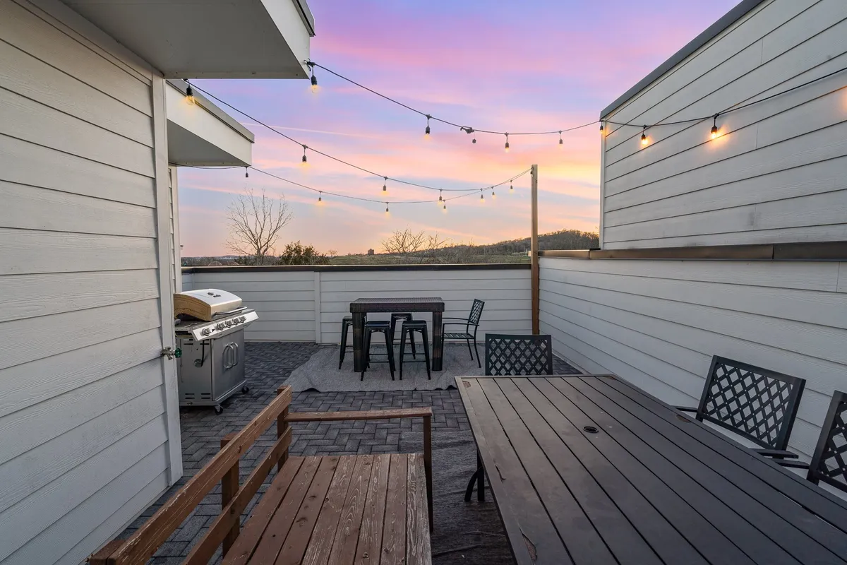 A rooftop patio scene featuring a sleek outdoor dining table, modern grill, patio furniture, and glowing string lights overhead. The space is arranged for easy socializing and outdoor meals, with the soft evening light enhancing its charm.